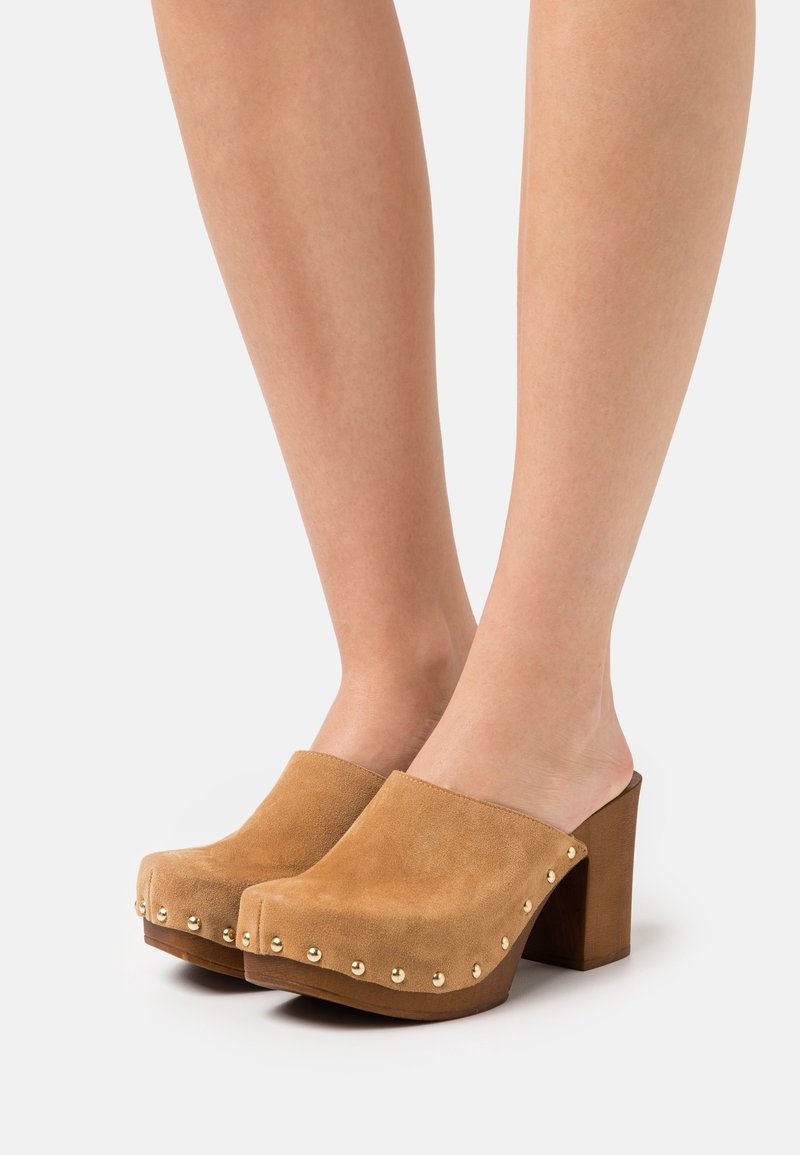 Sabots en cuir suédé beige avec un talon en bois, ornés d'oxydes métalliques sur le devant. Texture lisse et conception à dos ouvert.