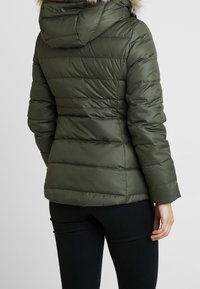 Veste matelassée vert olive à la silhouette cintrée, avec des coutures horizontales et une capuche bordée de fourrure. Texture lisse et manches longues.