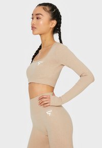 Bež kratki crop top s dugim rukavima, okruglim izrezom i rebrastom teksturom, u kombinaciji s hlačama visokog struka koje na kuku imaju bijeli logo.