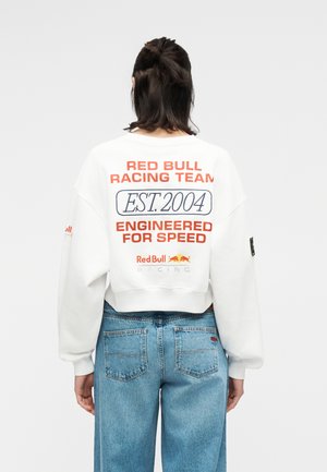 Persoană cu păr închis la culoare, purtând un hanorac alb Red Bull Racing Team cu text și blugi albaștri, stând cu spatele, pe un fundal simplu.