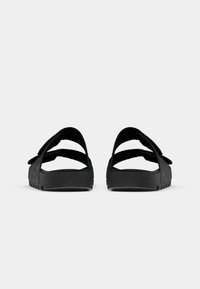 Et par svarte sandaler med åpen tå, med to justerbare stropper og teksturert såle, vist fra baksiden på en hvit bakgrunn.