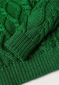 Manche de pull en maille torsadée verte et poignet côtelé pliés sur un fond de tissu texturé assorti.