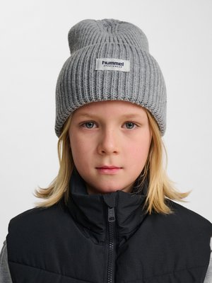 Dziecko z blond włosami noszące szarą prążkowaną dzianą czapkę z napisem „hummel sportswear” oraz czarną pikowaną kamizelkę na zamek.
