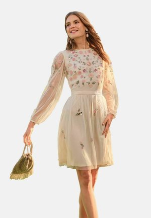 Žena nosí priehľadné šaty s dlhým rukávom s ružovým kvetinovým vyšívaním, drží malú textúrovanú kabelku, usmieva sa a kráča.