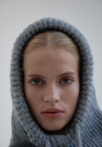 Szary dzianinowy hoodie o oversize'owym kroju z prążkowaną teksturą oraz dużym, zaokrąglonym kapturem, opadającym wokół twarzy.