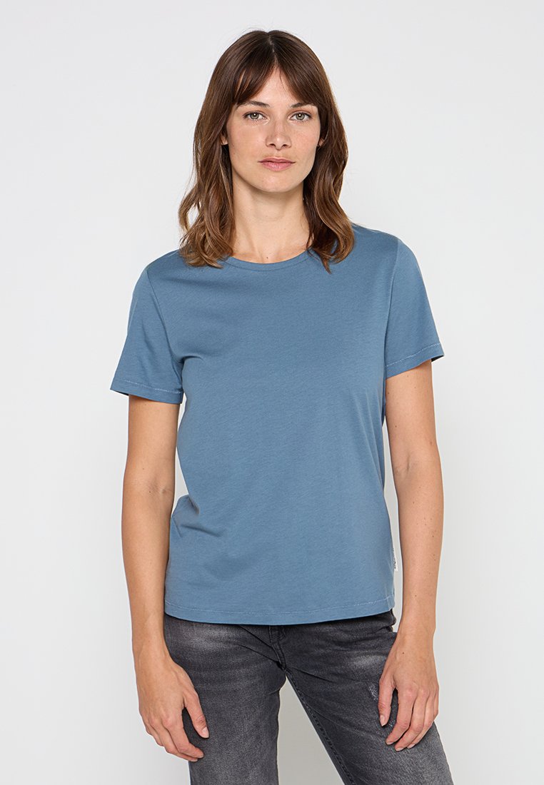 Blaues Baumwoll-T-Shirt mit rundem Halsausschnitt und kurzen Ärmeln, das eine glatte Textur und ein minimalistisches Design bietet. Kombiniert mit dunklen Jeans.