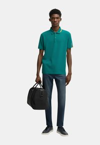 Polo teal con dettaglio giallo sul colletto, jeans blu scuro, borsa a tracolla nera e sneakers grigie, su una figura maschile su sfondo bianco.