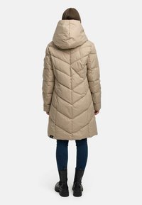 Beige puffjakke med stor hette, med et chevron kviltet mønster, som rekker til midten av låret, kombinert med mørke smale jeans og svarte støvler.