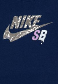 Tumsā zila T-kreklu rotā liels atstarots sudraba Nike logo ar „SB” purpursarkanā un baltā krāsā. Gluda kokvilnas tekstūra.