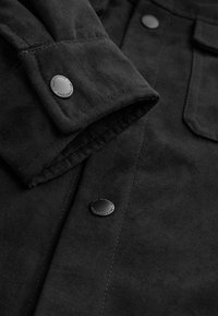 Schwarzes Wildledershirt mit umgeschlagenem Manschette und Druckknopf, glatte Textur und dezenten Glanz, mit sichtbaren Nähten und Taschenakzenten.