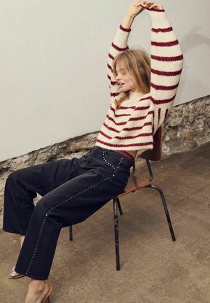 Striped wool sweater in cream and burgundy, paired with high-waisted black jeans featuring silver stud accents along the seams. Sitting on a chair.