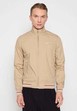 Beige leichte Jacke mit hohem Kragen, durchgehendem Reißverschluss und gerippten Bündchen. Mit roten und weißen Streifen am Saum und Markenlogo auf der Brust.
