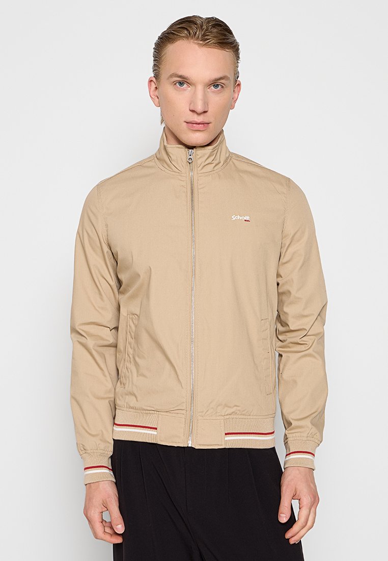 Veste légère beige avec un col montant, fermeture éclair intégrale et poignets côtelés. Comprend des accents rayés rouges et blancs au bas et un logo sur la poitrine.