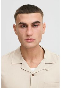 Beige kraagshirt van gladde stof, met een enkele knop, korte mouwen en een borstzak. Eenvoudig en modern design.