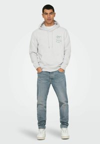 Sweatshirt gris avec une poche avant et un cordon de serrage, arborant un motif de palmier et du texte. Associé à un jean bleu clair et des baskets blanches.