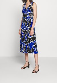 Sukienka bez rękawów w niebiesko-czarnym kolorze z wzorem łańcucha, z dekoltem w kształcie litery V, ściągniętym w talii i zwiewnym dołem. Czarne sandały na cienkiej szpilce.