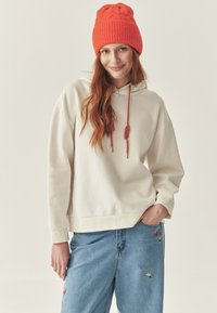 Sudadera blanca con cordón y detalles en rojo, combinada con unos vaqueros azul claro bordados con patrones florales y un gorro de punto naranja brillante.