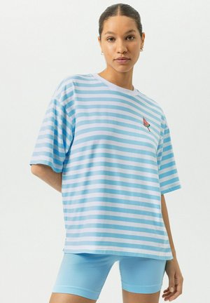 Femme portant un T-shirt ample à rayures bleu clair et blanc avec une petite écusson pastèque, et un short ajusté bleu clair, debout devant un fond blanc.