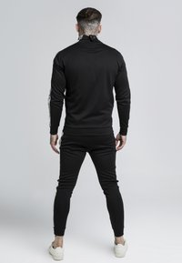 Black athletic outfit featuring a zip-up high neck and matching tapered pants. Details include white side stripes and ribbed cuffs.