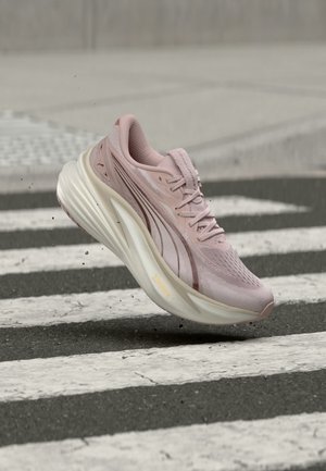 Lyserød atletiksneaker med tyk hvid sål svæver over et fodgængerfelt på en asfaltvej, små grusflager flyver rundt.