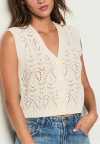 Pletená bezrukávová vesta v krémové barvě s výstřihem do V, zdobená krajkovým vzorem, žebrovanými okraji a pěti knoflíky vpředu. Nošena přes džínové kraťasy.