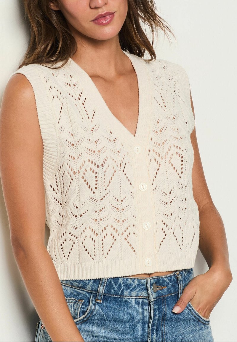 Pletená bezrukávová vesta v krémové barvě s výstřihem do V, zdobená krajkovým vzorem, žebrovanými okraji a pěti knoflíky vpředu. Nošena přes džínové kraťasy.