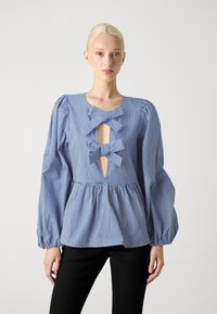 Μπλε και λευκή ριγέ μπλούζα με στυλ peplum, διαθέτοντας τρία διακοσμητικά φιόγκια και φουσκωτά μακριά μανίκια. Μαλακή υφή υφάσματος.