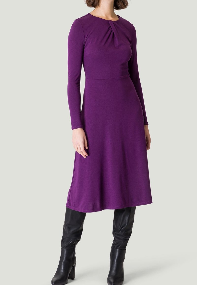 Robe violette à manches longues avec un col froncé, jupe fluide et texture lisse. Associée à des bottes noires à genoux.