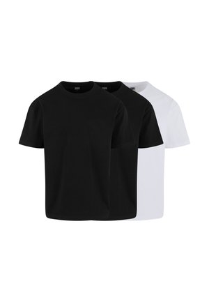 Jeune homme aux cheveux courts portant un t-shirt noir uni et un jean bleu clair, debout les mains dans les poches devant un fond gris.