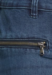 Tela de mezclilla azul con un detalle visible de cremallera metálica, que presenta un diseño de dientes dorados y un tirador plateado. Textura suave y forma recta.