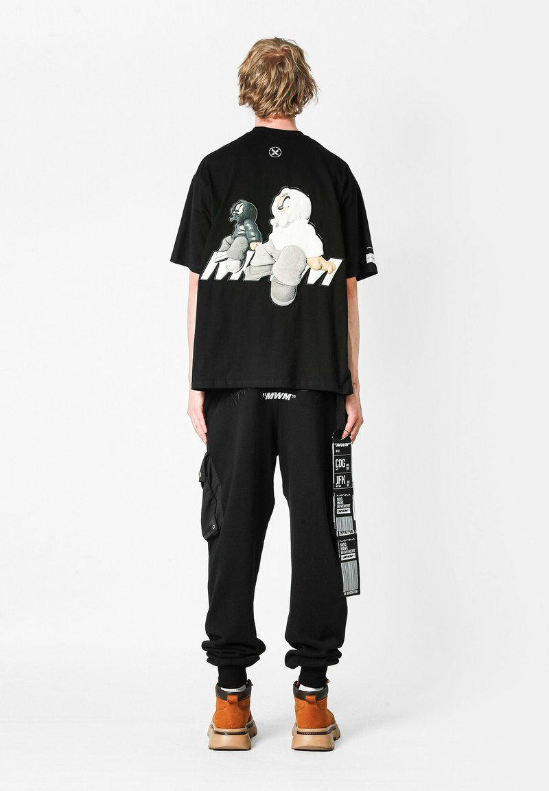 Camiseta negra de gran tamaño con estampado gráfico en la parte posterior, combinada con pantalones jogger negros con bolsillos de carga y acentos de texto en blanco. Botas marrones.