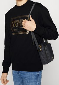 Svart sweatshirt med guld-logotyp, avslappnad passform. Par av ljusblå slitna jeans. Liten grå axelväska med rem och guldaccenter.