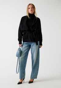 Sort strikket cardigan med sjalkrave, bælte i taljen, parret med lyseblå hochwaisted jeans og sorte spidse hæle, mens han holder en lyseblå taske.