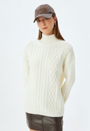 Maglione crema a trecce con collo alto e vestibilità ampia, abbinato a un berretto di pelle marrone e a una gonna di pelle marrone scuro.