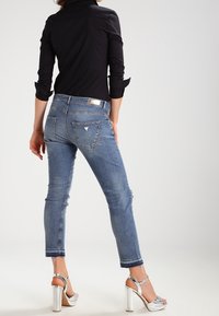 Mujer vestida con jeans azules con puños, camiseta negra de manga larga y sandalias plateadas de tacón alto, de pie frente a un fondo liso.