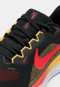 Chaussure de course Nike noire et jaune avec logo swoosh rouge et lacets noirs, montrant des coutures détaillées et un tissu mesh texturé.