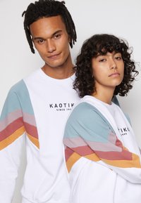 Hvid sweatshirt med et multicolor diagonalt stribedesign i blå, pink og orange. Brandlogo vist centralt. Materialet er en blanding af bomuld.