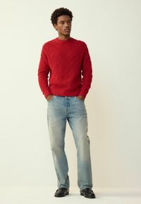 Piros, texturált kötött pulóver kerek nyakkivágással, világoskék kifakult, egyenes szárú farmerral és fekete mokaszinokkal, semleges háttér előtt állva.