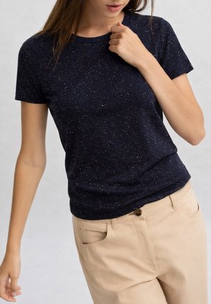 Donna che indossa una maglietta a maniche corte blu scuro maculata e pantaloni beige, con una mano vicino al colletto e l'altra lungo il fianco.