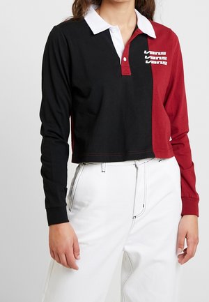 Kobieta nosząca długi rękaw, koszulkę polo z blokiem kolorów w czerni i czerwieni z białym kołnierzem i logo Vans, zestawioną z białymi spodniami.