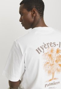 Witte katoenen t-shirt met een grote oranje palmboomgrafiek op de achterkant en de tekst "Hyères-Les-Palmiers" erboven. Korte mouwen, losse pasvorm.
