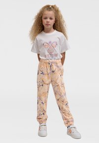 Weißes T-Shirt mit "CULTURE" Grafik, kombiniert mit orangefarbenen, abstrakt gemusterten Sweathosen, die einen elastischen Bund und Bündchen besitzen. Weiße Schuhe.