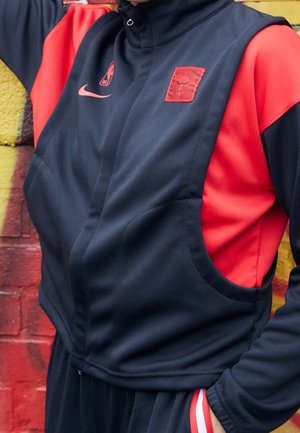 Person trägt eine schwarz-rote Jacke mit Nike-Logo und rotem Emblem, steht vor einer bunt bemalten Ziegelwand.
