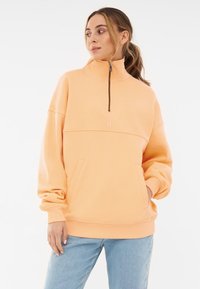 Licht perzikkleurige kwart-rits pullover met een losse pasvorm, een kangoeroezak en geribde manchetten. Zachte stof met subtiele logodetails.