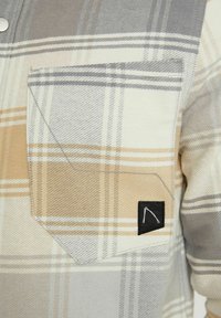Gros plan d'une poche de chemise à carreaux beige, gris et blanc avec une petite étiquette noire rectangulaire affichant un motif simple en lignes blanches.