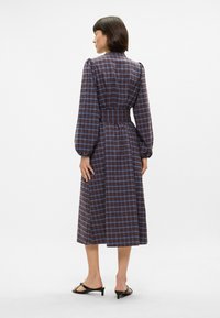 Langermet, rutete kjole i nyanser av brunt og blått, med tilpasset midje og løs skjørt. Glatt stoff, knyttet krage og puffede ermer.