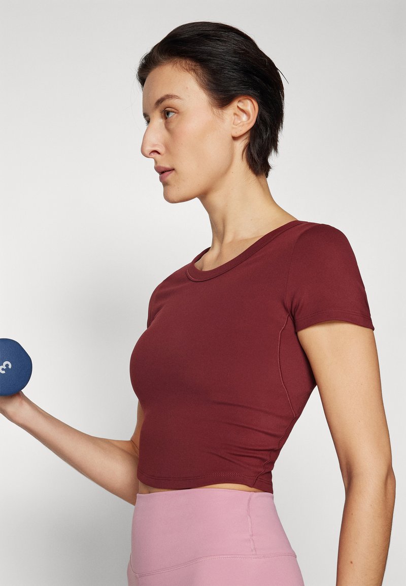 Crop top bordeaux con maniche corte, texture liscia e scollatura tonda, abbinato a leggins rosa a vita alta e un manubrio blu.