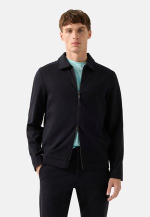 Mladý muž v černé bundě na zip přes světle modrou košili, stojící s rukama v kapsách před jednobarevným pozadím.