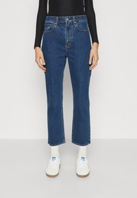 Jeans in denim blu con vita alta e vestibilità dritta, caratterizzati da un classico design a cinque tasche e cuciture a contrasto. Abbinati a sneakers bianche.