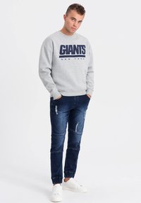 Sweatshirt gris avec "GIANTS NEW YORK" imprimé en bleu marine, associé à un jean bleu foncé avec des détails déchirés et des baskets blanches.